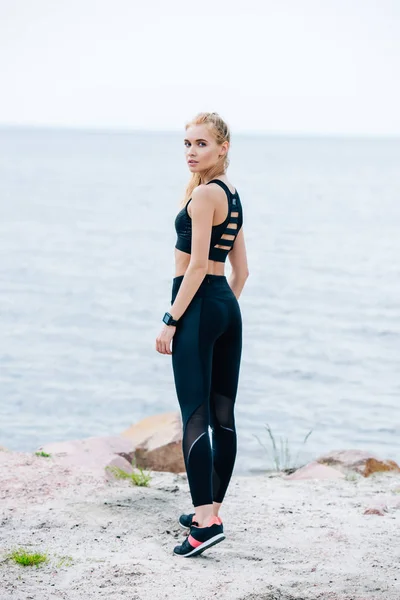 Attrayant blonde jeune femme en vêtements de sport regardant la caméra tout en se tenant près de la mer — Photo de stock