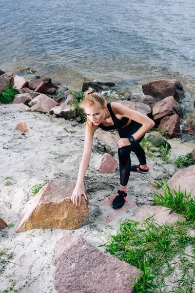 Overhead view of beautiful blonde sportswoman in sportswear climbing near stones and sea — Stock Photo