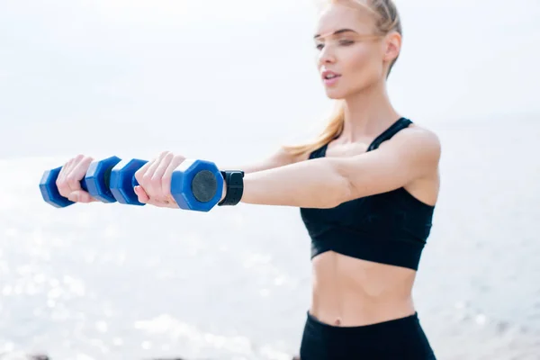 Selektiver Fokus des blonden athletischen Mädchens, das in der Nähe des Meeres mit Kurzhanteln trainiert — Stockfoto