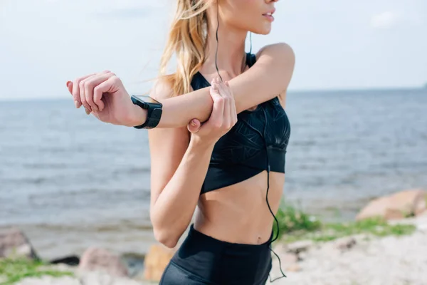 Cropped view of athletic girl stretching and listening music in earphones near sea — Stock Photo