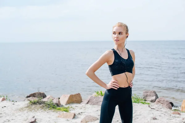 Mujer atlética de pie con las manos en las caderas y escuchar música en los auriculares cerca del mar - foto de stock