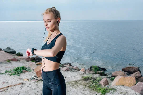 Atractiva mujer rubia mirando el rastreador de fitness y escuchando música en los auriculares cerca del mar - foto de stock