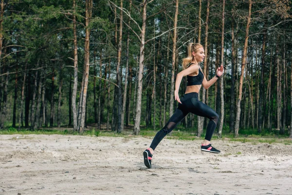 Счастливая и спортивная блондинка, бегущая возле зеленых деревьев — стоковое фото