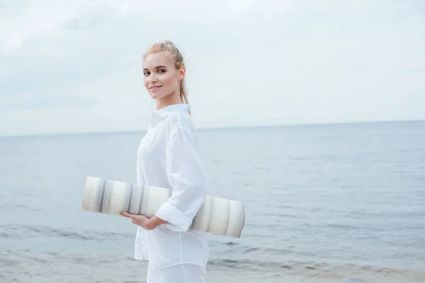 Attractive blonde woman smiling while holding yoga mat and standing near sea — Stock Photo