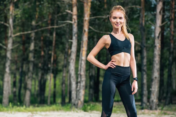 Glückliche athletische Frau, die mit der Hand auf der Hüfte steht und in die Kamera blickt — Stockfoto