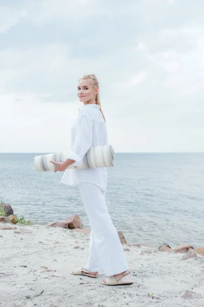 Sorridente donna bionda in possesso di tappetino yoga e in piedi vicino al mare — Foto stock