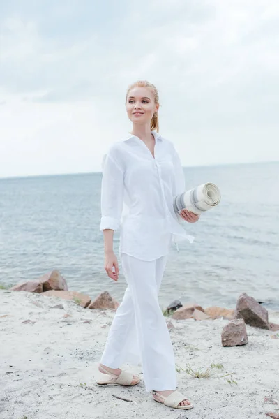 Счастливая блондинка, держащая коврик для йоги и стоящая у моря — стоковое фото