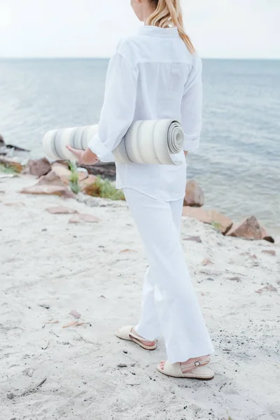Vista recortada de la joven mujer sosteniendo estera de yoga y de pie cerca del mar - foto de stock