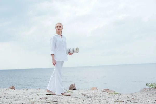 Glückliche junge blonde Frau, die am Meer steht und eine Yogamatte in der Hand hält — Stockfoto