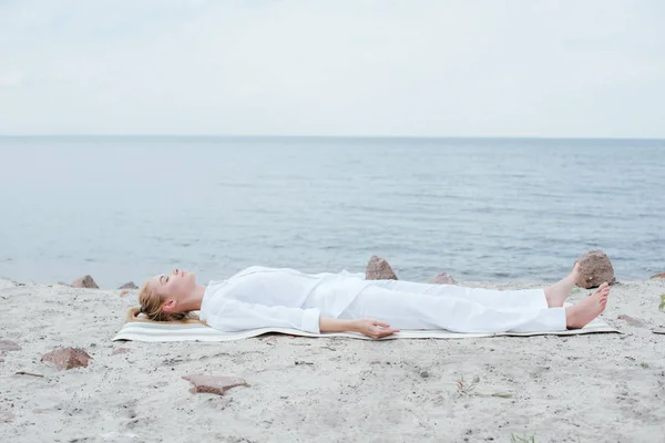 Mulher loira atraente com olhos fechados meditando enquanto deitado no tapete de ioga perto do mar — Fotografia de Stock