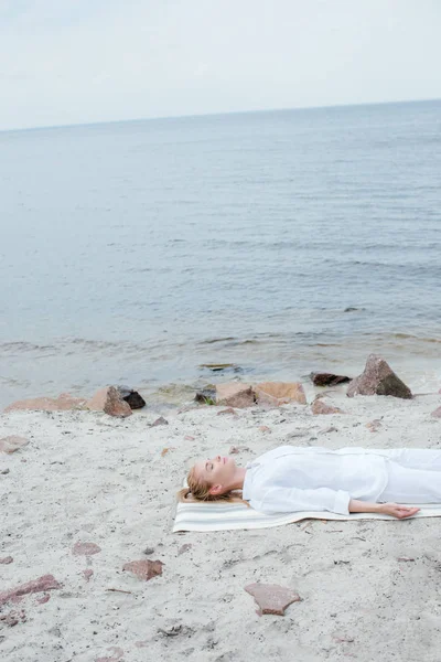 Belle femme blonde avec les yeux fermés méditant tout en étant couché sur un tapis de yoga près de la mer — Photo de stock