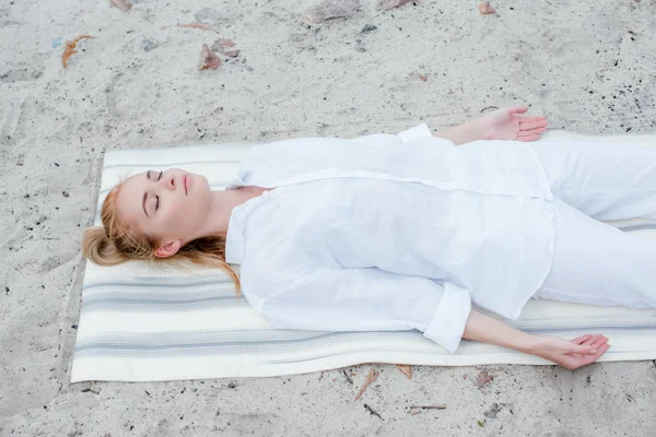 Overhead view of attractive blonde woman with closed eyes meditating while lying on yoga mat — Stock Photo