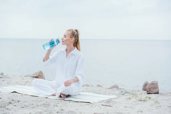 Attraktive Frau sitzt auf Yogamatte und hält Sportflasche, während sie Wasser trinkt — Stockfoto