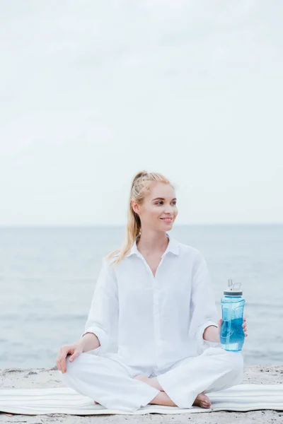 Mujer rubia alegre sosteniendo la botella del deporte y sentado en la estera del yoga cerca del mar - foto de stock