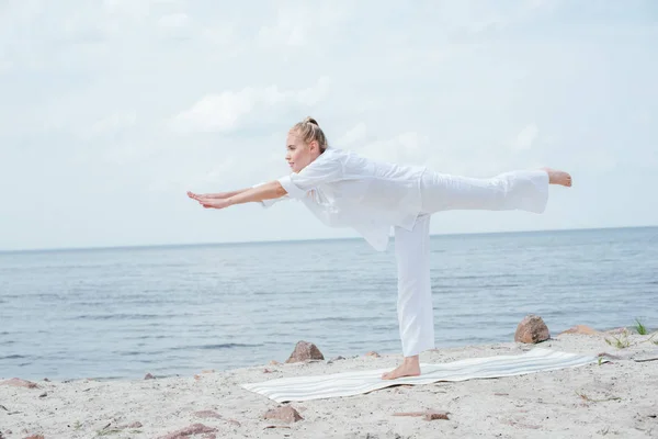 Attractive blonde woman practicing yoga on yoga mat near sea — Stock Photo