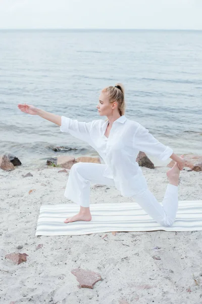 Vista lateral de atractiva mujer rubia practicando yoga en estera de yoga cerca del mar - foto de stock