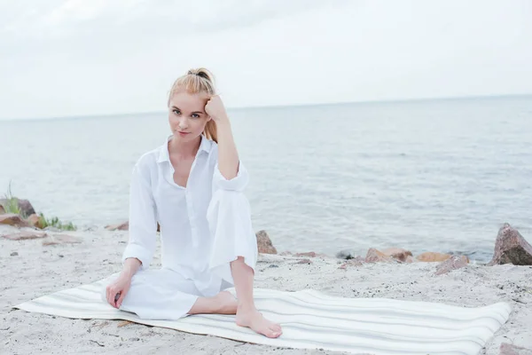 Bella giovane donna seduta su tappetino yoga e guardando la fotocamera vicino al mare — Foto stock