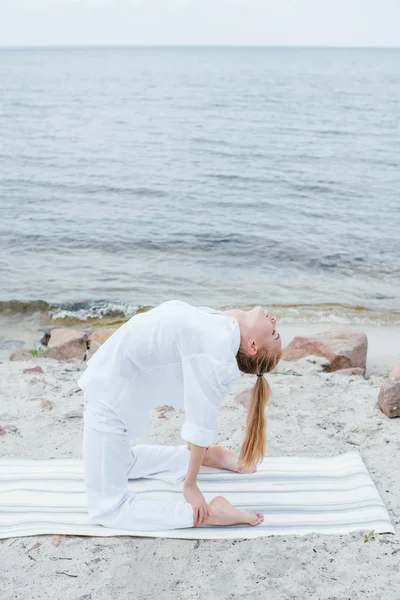 Rubia joven con los ojos cerrados practicando yoga en la esterilla de yoga cerca del mar - foto de stock