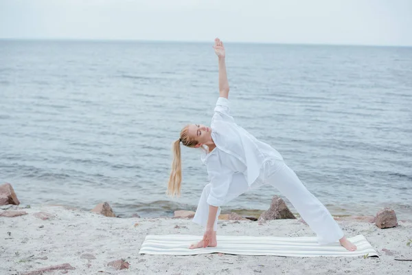 Chica atractiva con los ojos cerrados practicando yoga en la esterilla de yoga cerca del mar - foto de stock