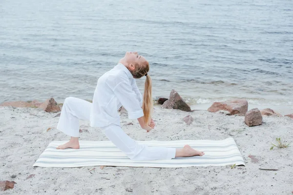 Hermosa chica con los ojos cerrados practicando yoga en estera de yoga cerca del mar - foto de stock