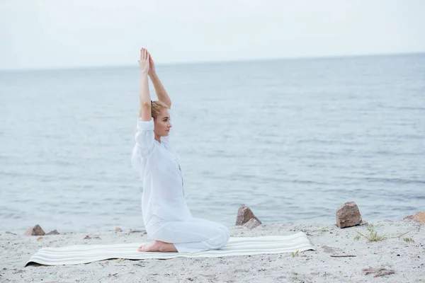 Atractiva mujer rubia practicando yoga con las manos sobre la cabeza cerca del río - foto de stock
