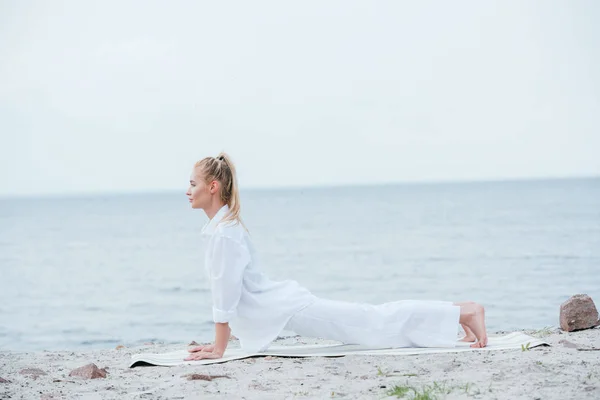 Vista lateral de la joven rubia practicando yoga cerca del río en la esterilla de yoga - foto de stock