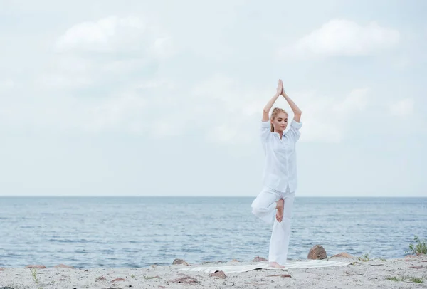 Attractive blonde woman practicing yoga with hands above head near sea — Stock Photo