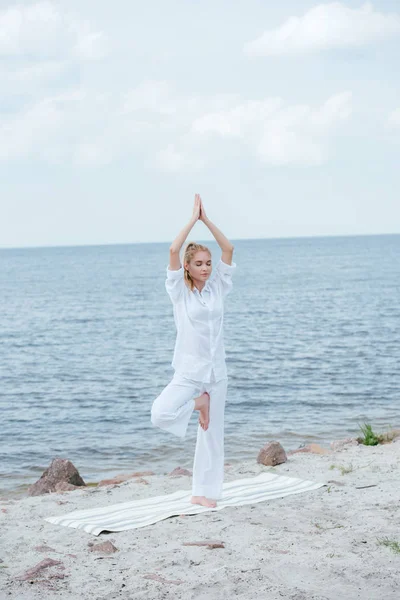 Atractiva mujer rubia practicando yoga con las manos por encima de la cabeza en la esterilla de yoga - foto de stock