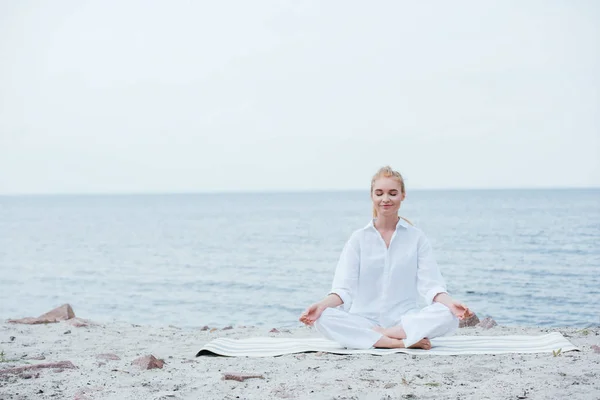 Счастливая блондинка с закрытыми глазами сидит на коврике для йоги — стоковое фото
