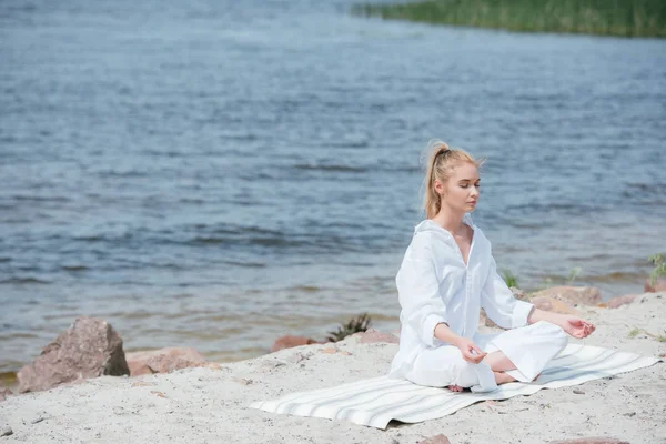 Миролюбивая блондинка с закрытыми глазами практикующая йогу возле реки — стоковое фото