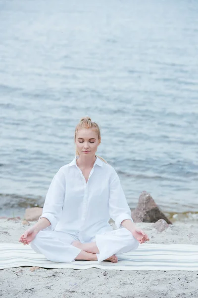 Paisible jeune femme blonde aux yeux fermés pratiquant le yoga près de la rivière — Photo de stock