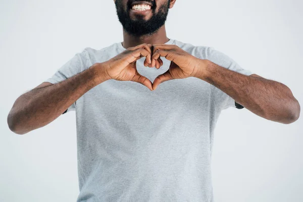 Abgeschnittene Ansicht eines lächelnden afrikanisch-amerikanischen Mannes in grauem T-Shirt, das Herzzeichen isoliert auf grau zeigt — Stockfoto