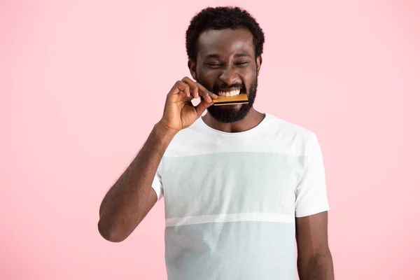 Hombre afroamericano mordiendo tarjeta de crédito aislado en rosa - foto de stock