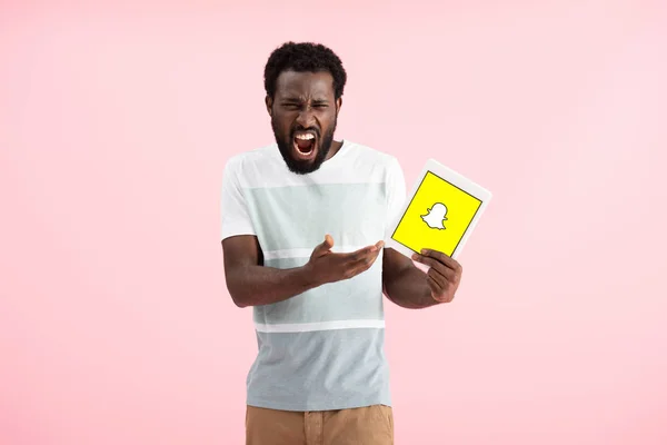 KYIV, UKRAINE - MAY 17, 2019: emotional african american man shouting and showing digital tablet with Snapchat app, isolated on pink — Stock Photo