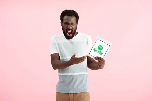 KYIV, UCRANIA - 17 DE MAYO DE 2019: hombre afroamericano emocional gritando y mostrando tableta digital con aplicación spotify, aislado en rosa - foto de stock