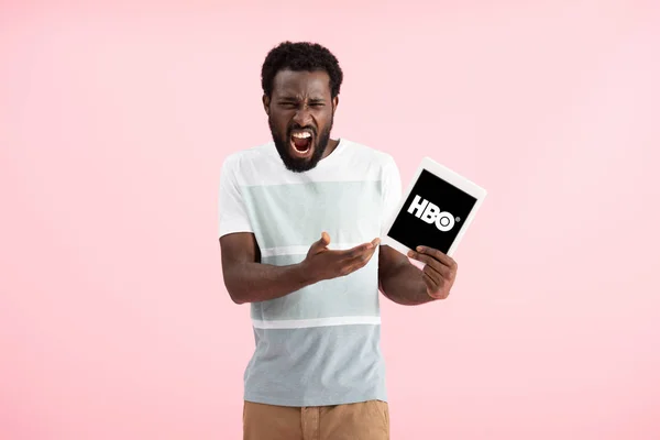 KYIV, UKRAINE - 17 MAI 2019 : un homme afro-américain émotionnel criant et montrant une tablette numérique avec l'application HBO, isolé sur rose — Photo de stock