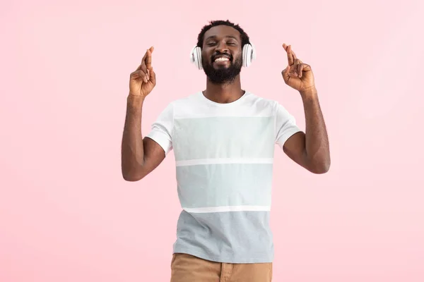 Feliz afro-americano com olhos fechados e dedos cruzados ouvindo música com fones de ouvido, isolado em rosa — Fotografia de Stock