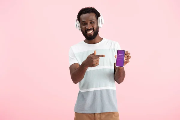 KYIV, UKRAINE - MAY 17, 2019: african american man listening music with headphones and pointing at smartphone with instagram app, isolated on pink — Stock Photo