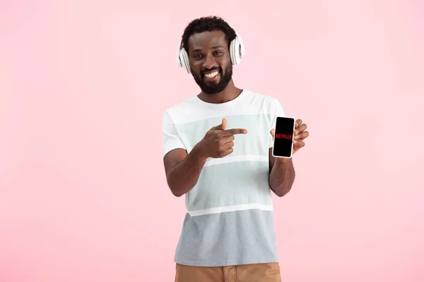 Kyiv, ukraine - 17. Mai 2019: afrikanisch-amerikanischer Mann hört Musik mit Kopfhörern und zeigt auf Smartphone mit Netflix-App, isoliert auf rosa — Stockfoto