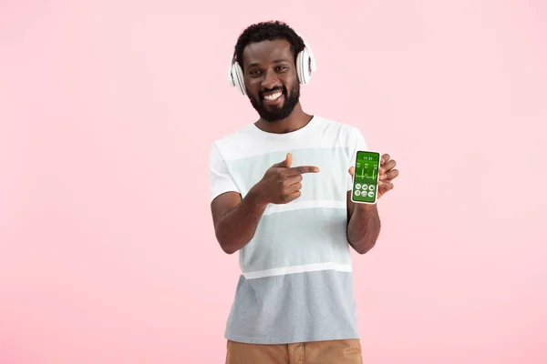 Hombre afroamericano escuchando música con auriculares y apuntando al teléfono inteligente con aplicación de salud, aislado en rosa - foto de stock