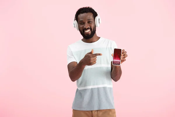 Afro-americano ouvindo música com fones de ouvido e apontando para smartphone com cursos comerciais, isolado em rosa — Fotografia de Stock