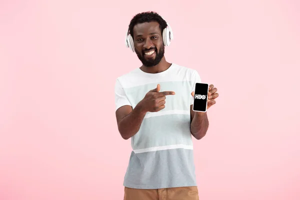 KYIV, UCRAINA - 17 MAGGIO 2019: uomo afroamericano che ascolta musica con le cuffie e punta lo smartphone con l'app HBO, isolato su rosa — Foto stock