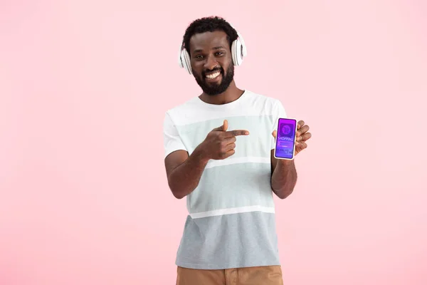Afrikanisch-amerikanischer Mann hört Musik mit Kopfhörern und zeigt auf Smartphone mit Shopping-App, isoliert auf rosa — Stockfoto
