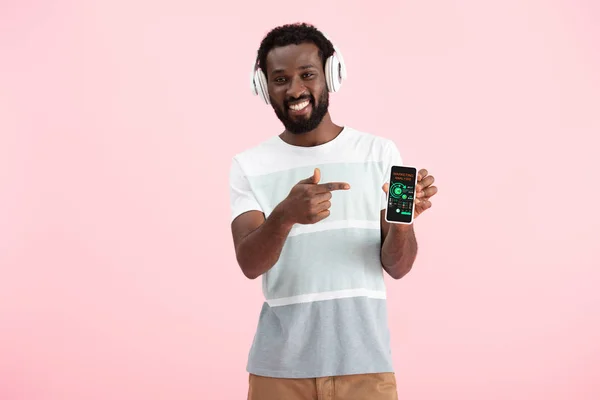 Afrikanisch-amerikanischer Mann hört Musik mit Kopfhörern und zeigt auf Smartphone mit Marketing-Analyse, isoliert auf rosa — Stockfoto