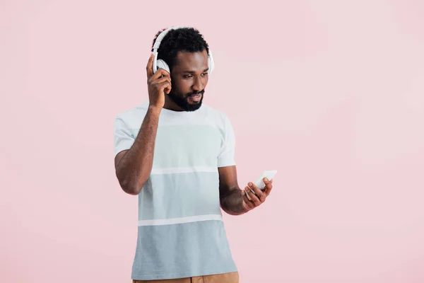 Hombre afroamericano escuchando música con auriculares y smartphone, aislado en rosa - foto de stock