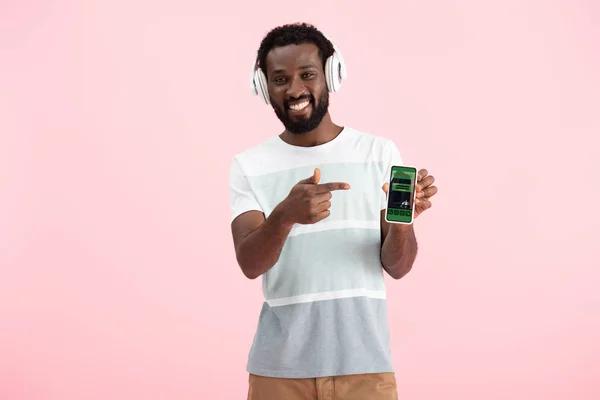 Afrikanisch-amerikanischer Mann hört Musik mit Kopfhörern und zeigt auf Smartphone mit Buchungs-App, isoliert auf rosa — Stockfoto