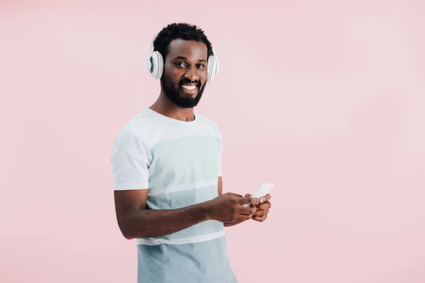 Homem americano africano alegre ouvindo música com fones de ouvido e smartphone, isolado em rosa — Fotografia de Stock