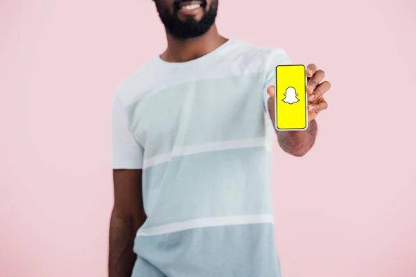 KYIV, UCRANIA - 17 de mayo de 2019: vista recortada del hombre afroamericano que muestra el teléfono inteligente con la aplicación Snapchat, aislado en rosa - foto de stock