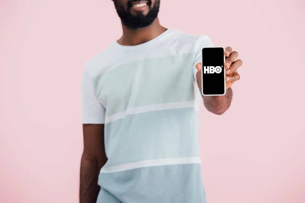 KYIV, UCRANIA - 17 de mayo de 2019: vista recortada del hombre afroamericano mostrando su teléfono inteligente con la aplicación HBO, aislado en rosa - foto de stock