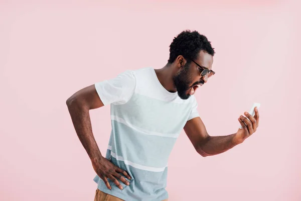 Aggressive african american of man in sunglasses shouting at smartphone, isolated on pink — Stock Photo